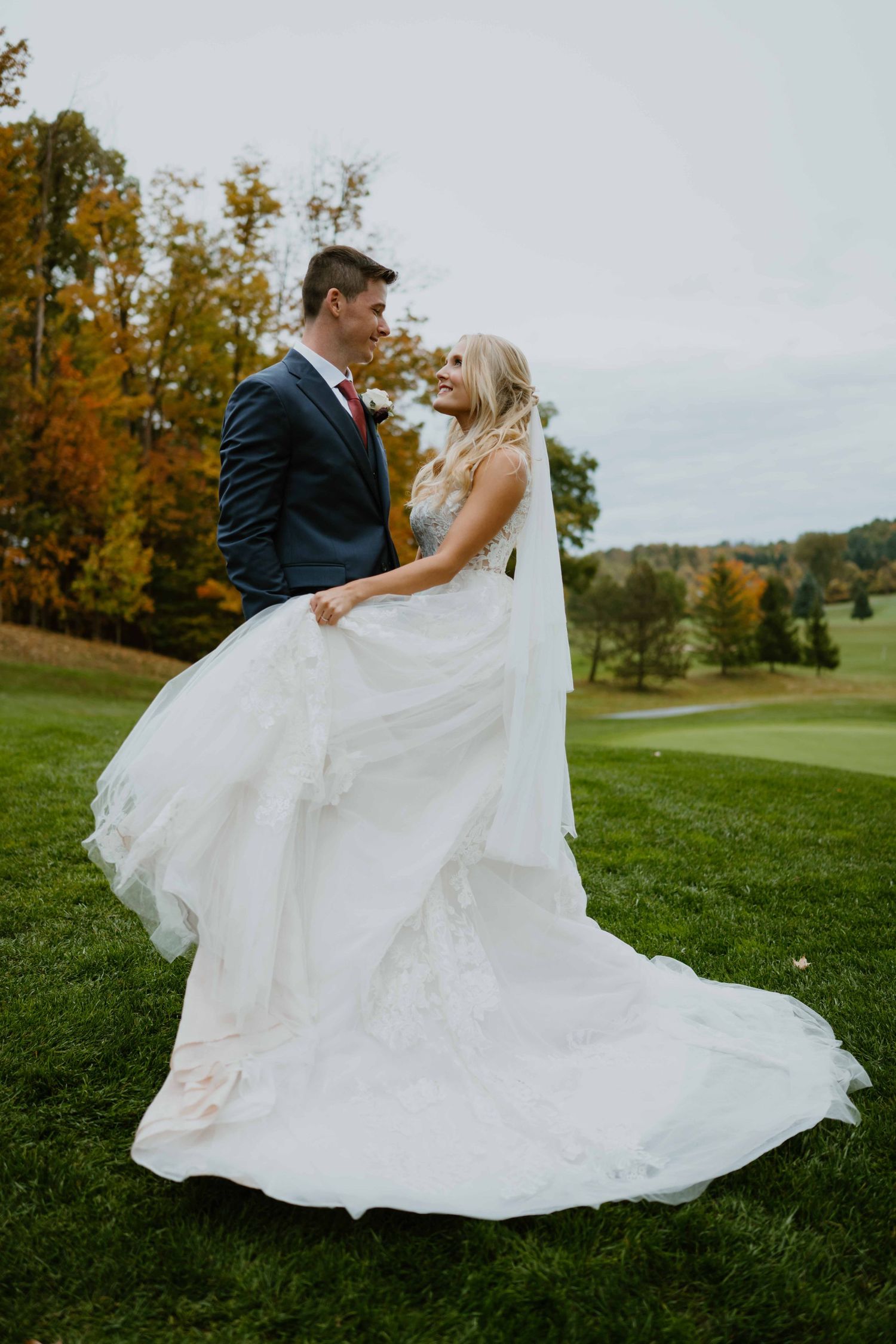 Groom twirling bride in flowing white gown on green golf course in fall.