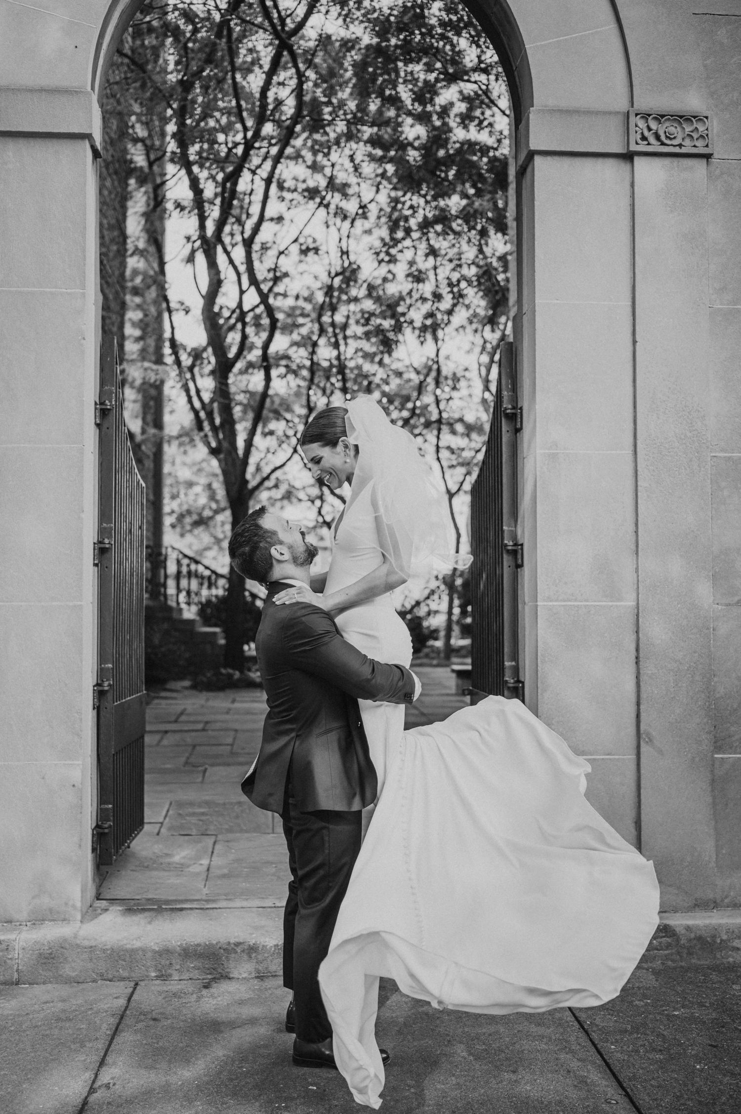 Black and white photo of groom lifting bride under arched doorway surrounded by trees.