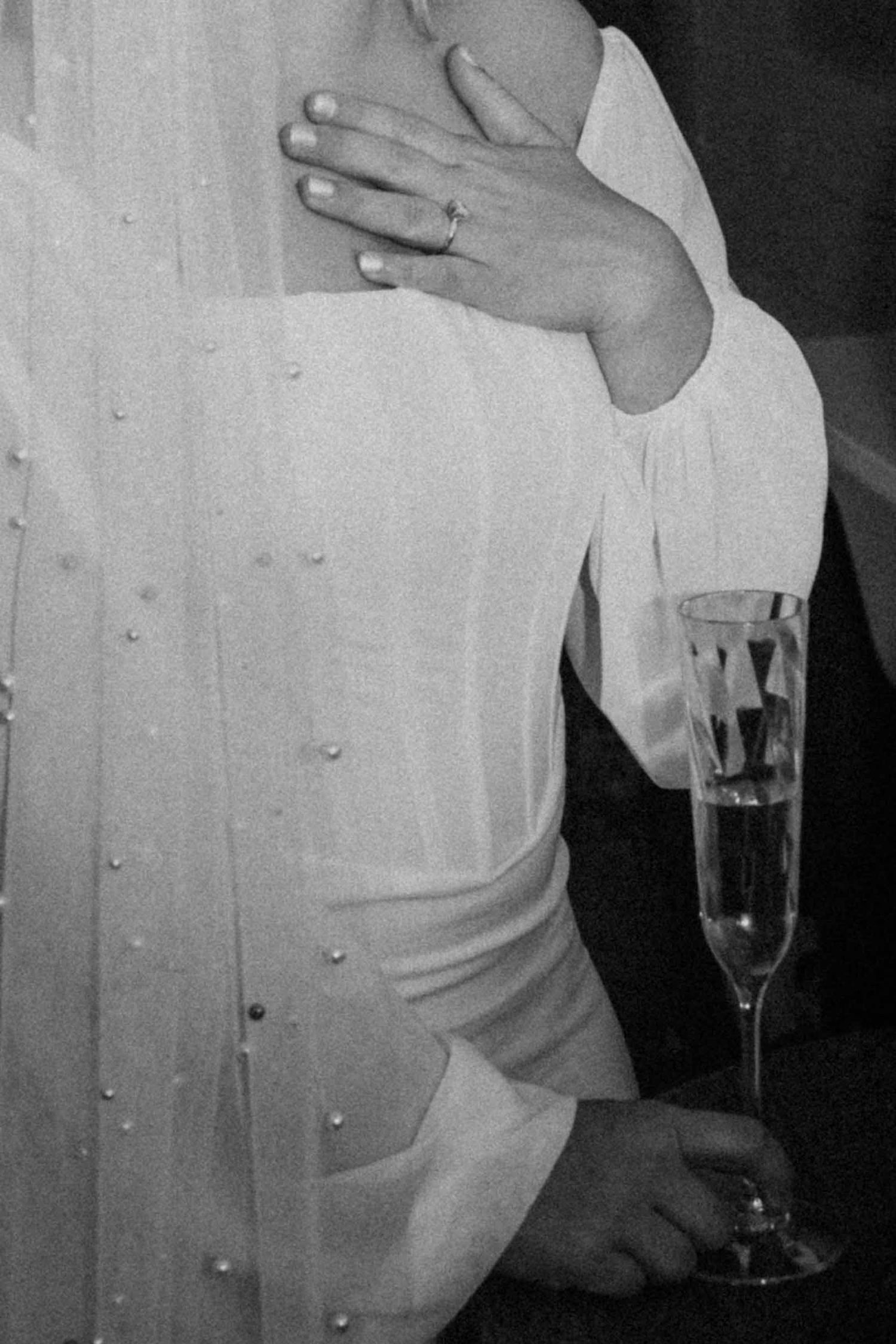 Black and white close-up of bride in pearl veil holding champagne flute with wedding ring visible.
