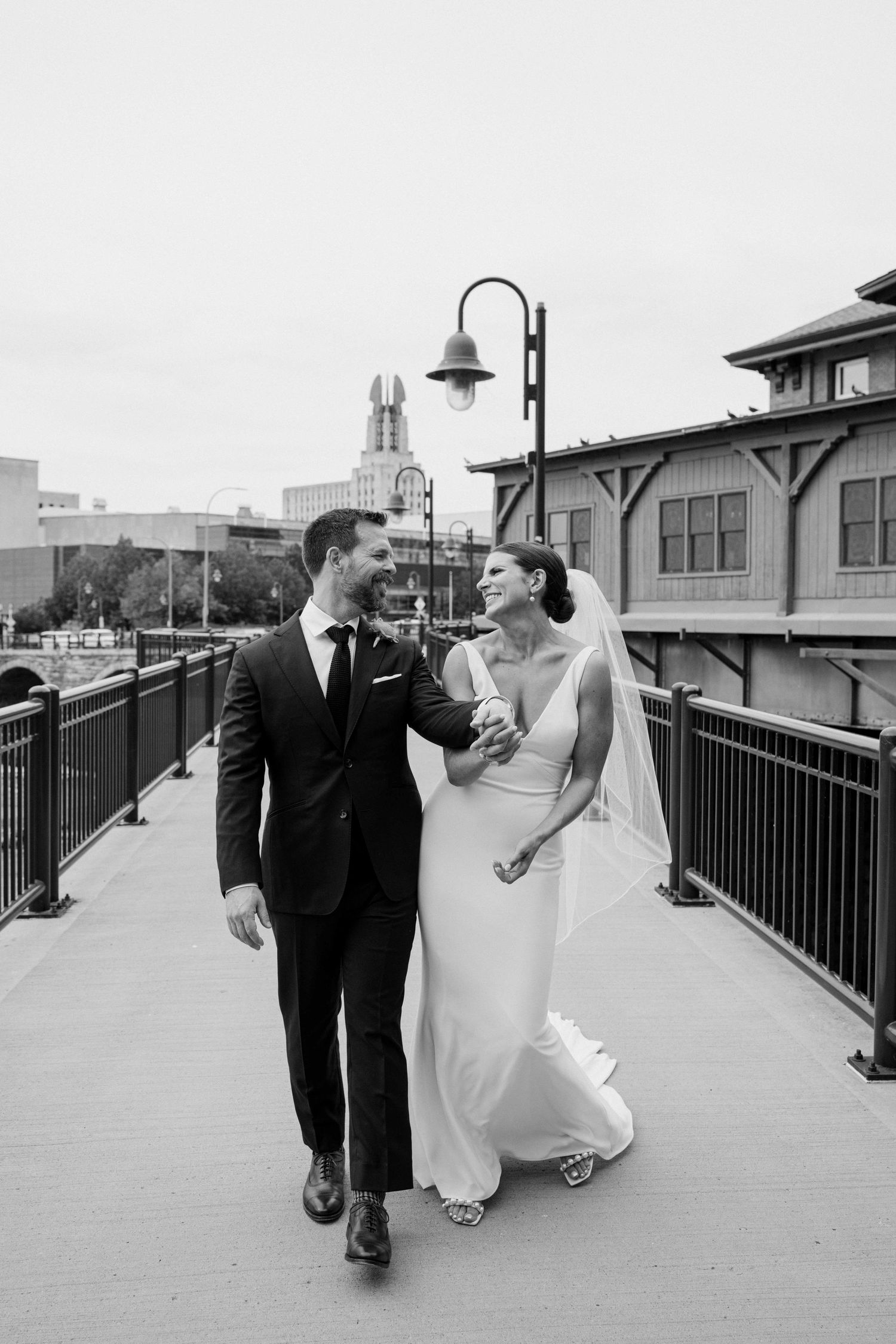 Bride and groom walk together on urban bridge, smiling, in black and white wedding portrait.