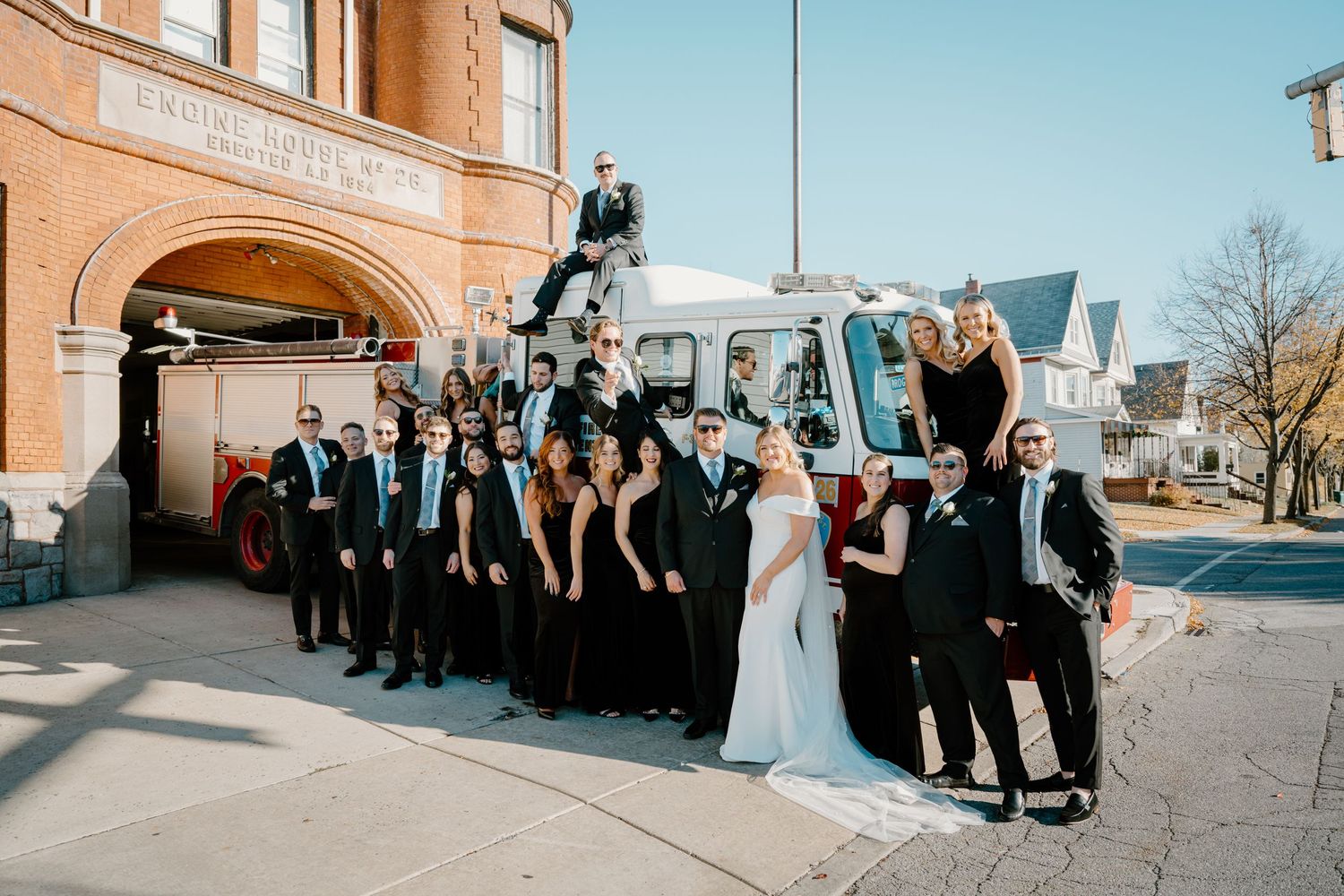 Wedding party poses with vintage fire truck outside historic brick engine house on sunny day.