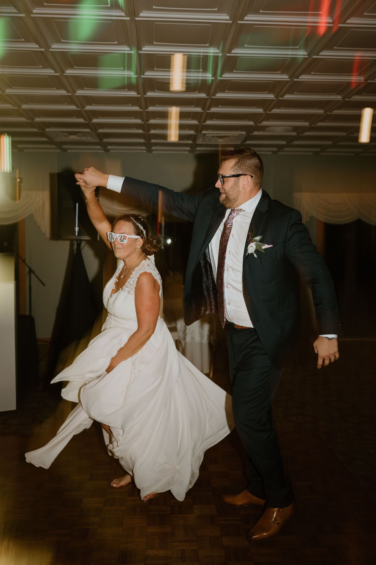 Bride wearing fun glasses being twirled by groom during energetic wedding reception dance celebration.