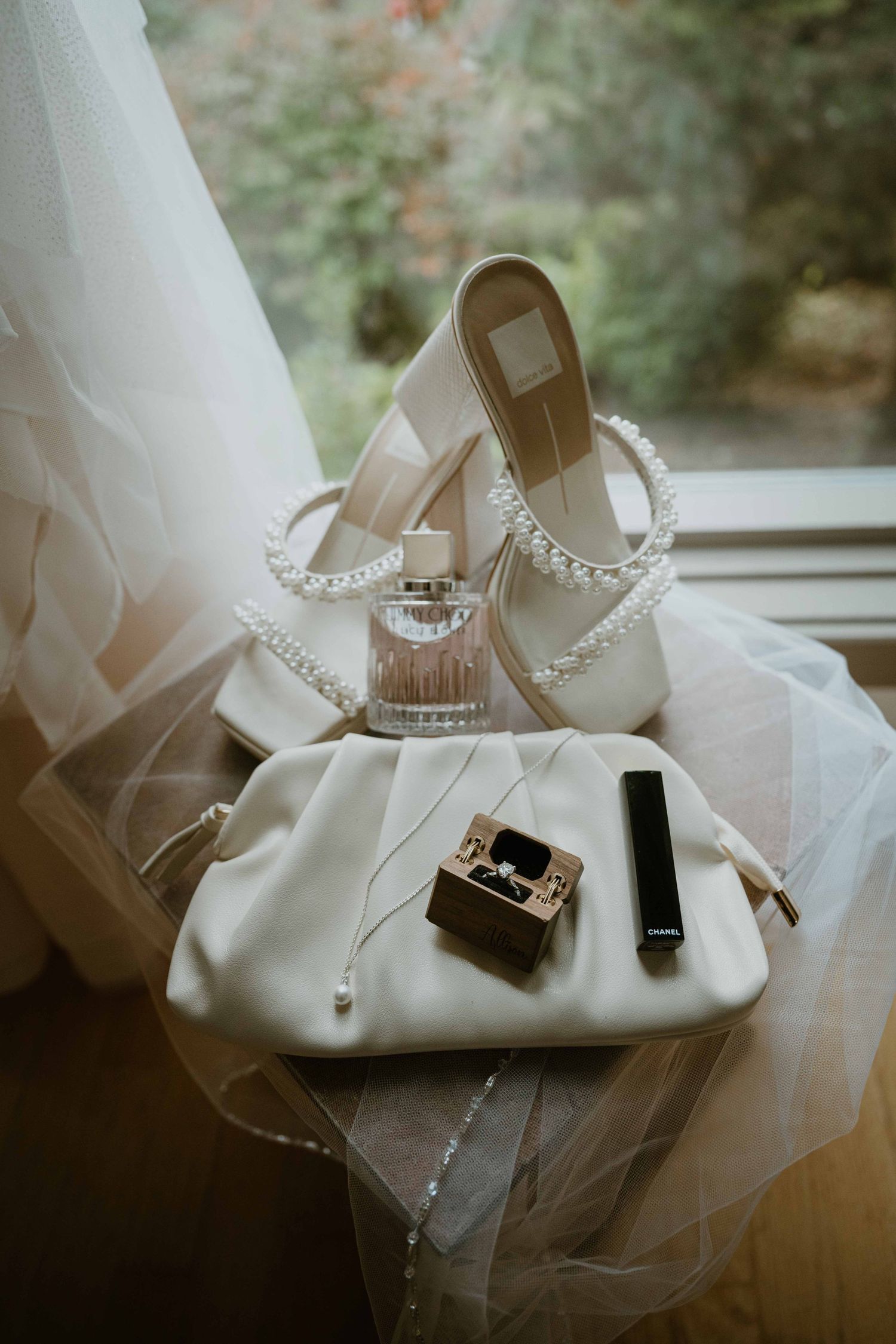 Bridal accessories flatlay featuring pearl heels, white clutch, perfume, ring box by window.