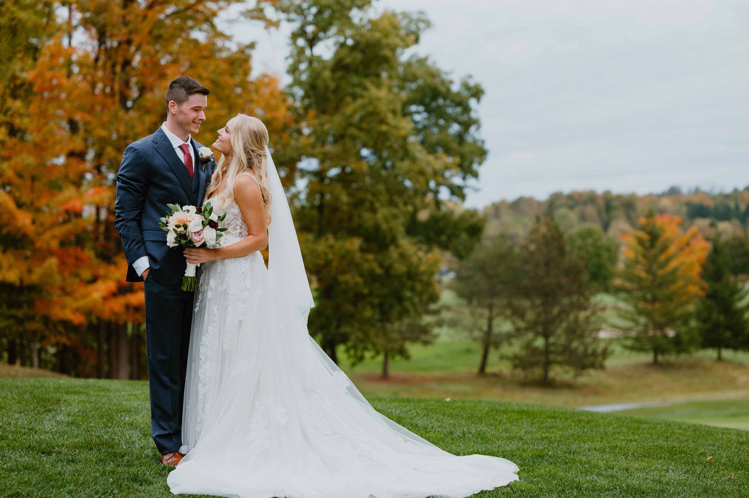 Bride and groom posing on golf course surrounded by vibrant fall foliage autumn wedding.