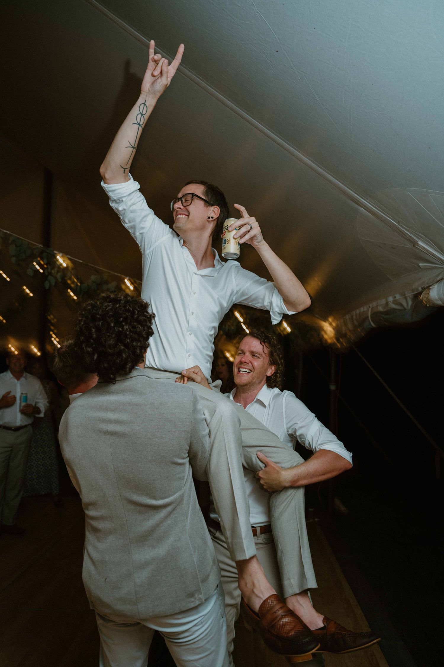 Groomsman lifted on shoulders celebrates joyfully at lively indoor wedding reception party.