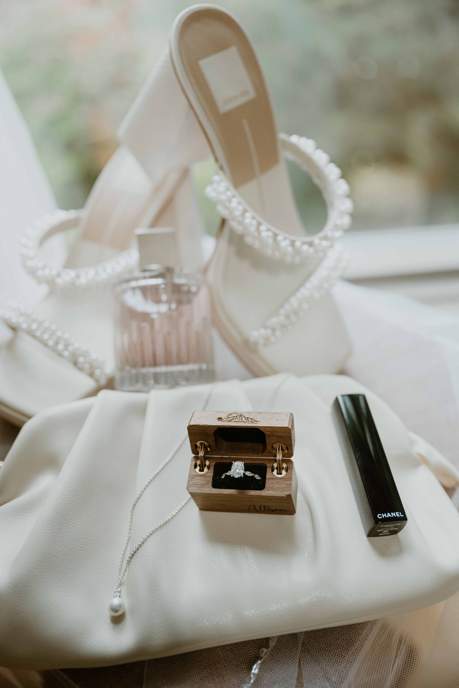 Wedding detail flatlay with diamond ring in wooden box, Chanel mascara, pearl heels, perfume.
