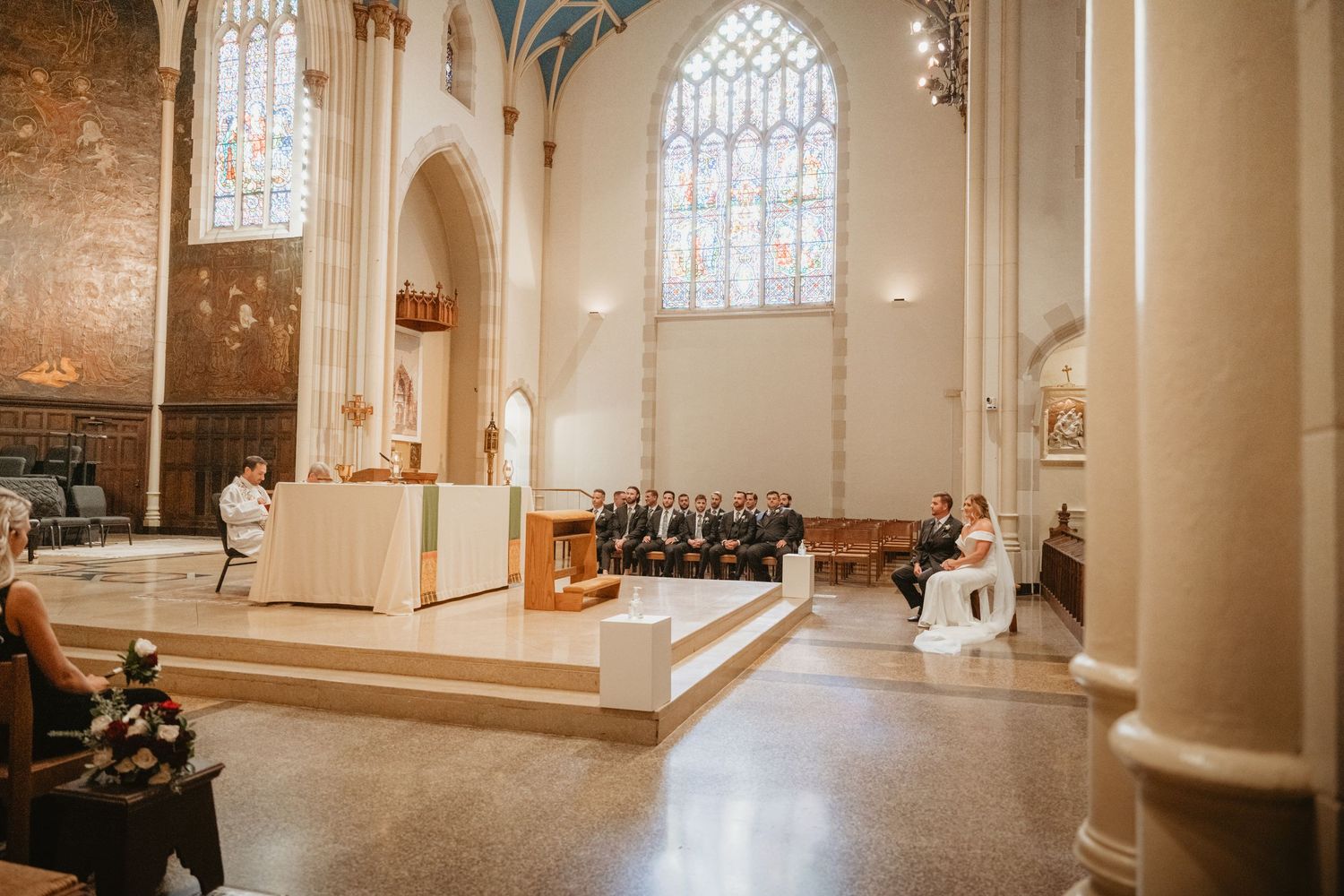 Elegant church wedding ceremony with bride and groom kneeling at altar beneath stunning stained glass windows.