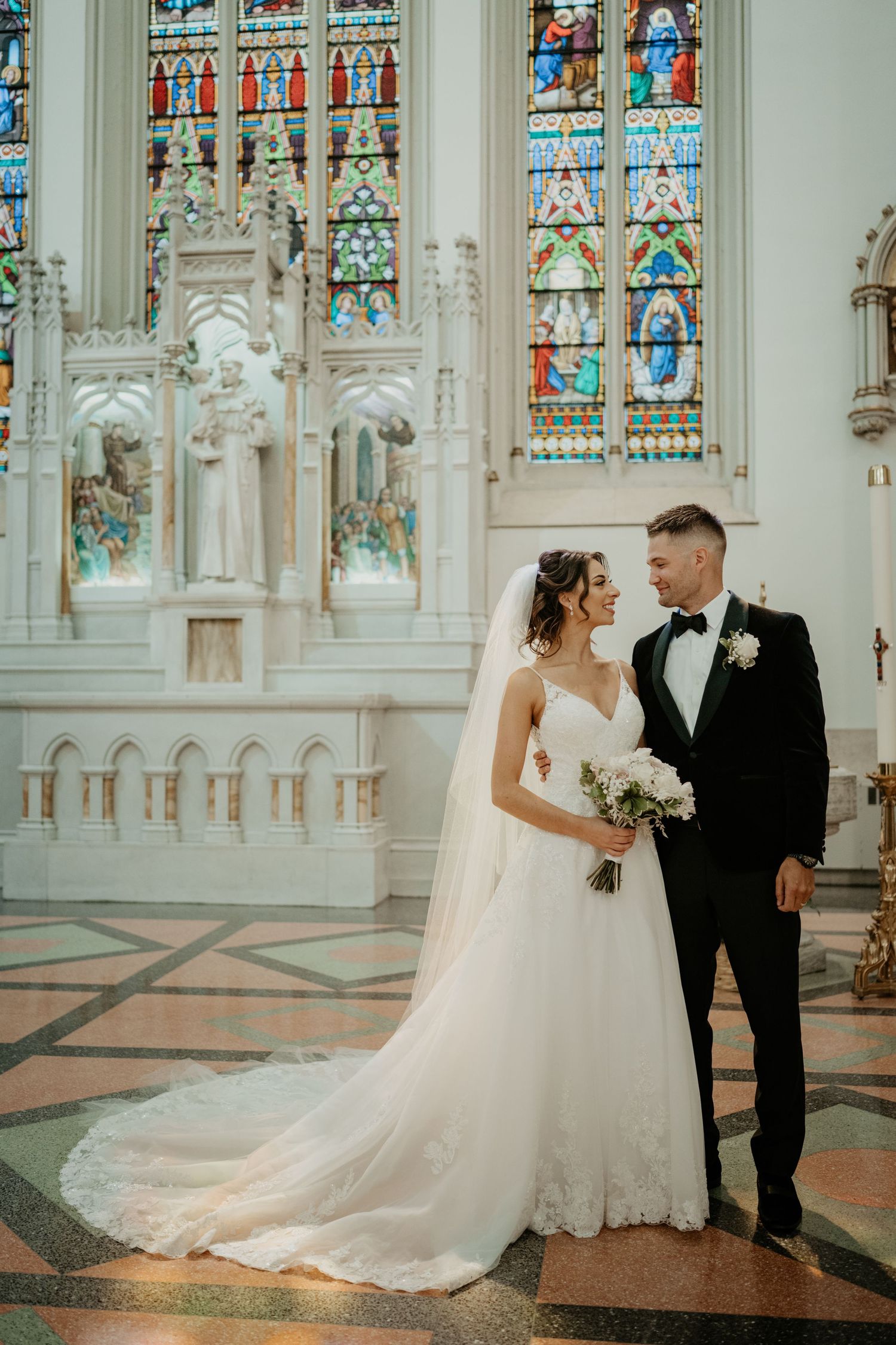 Elegant bride and groom portrait inside ornate Gothic church with colorful stained glass windows backdrop.