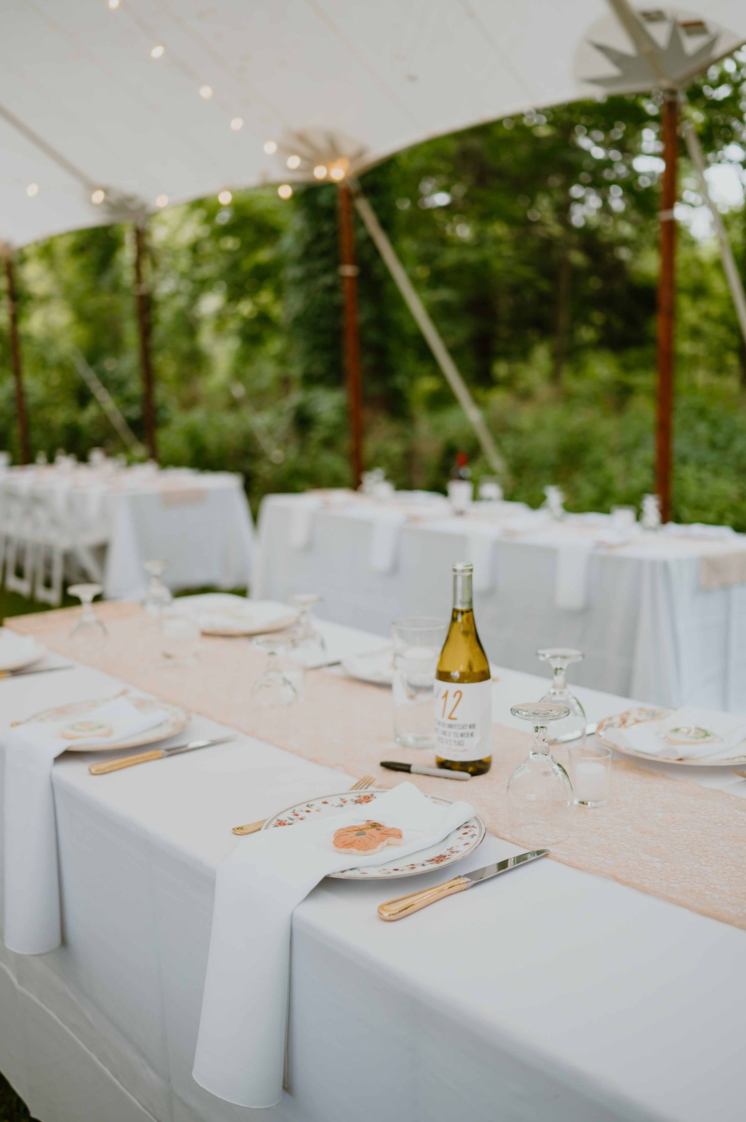 Elegant outdoor wedding reception tables set with white linens, blush runners, and white wine under a tent.