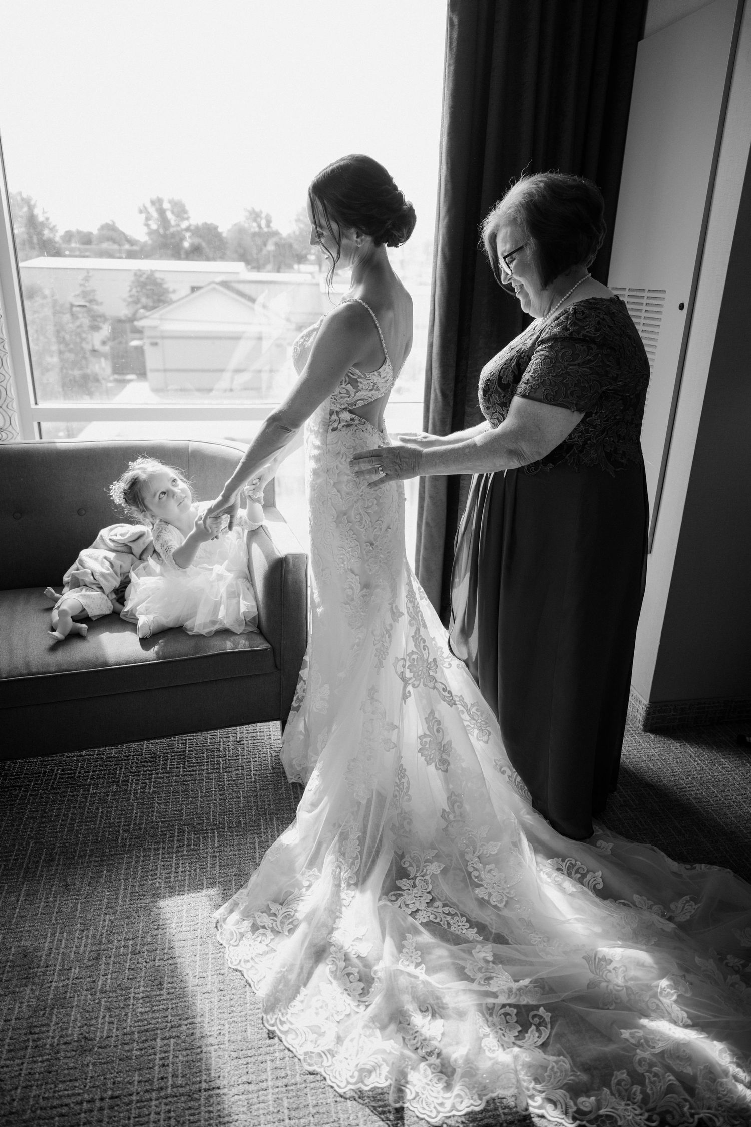 Black and white photo of mother helping bride into her lace wedding gown by a bright window.