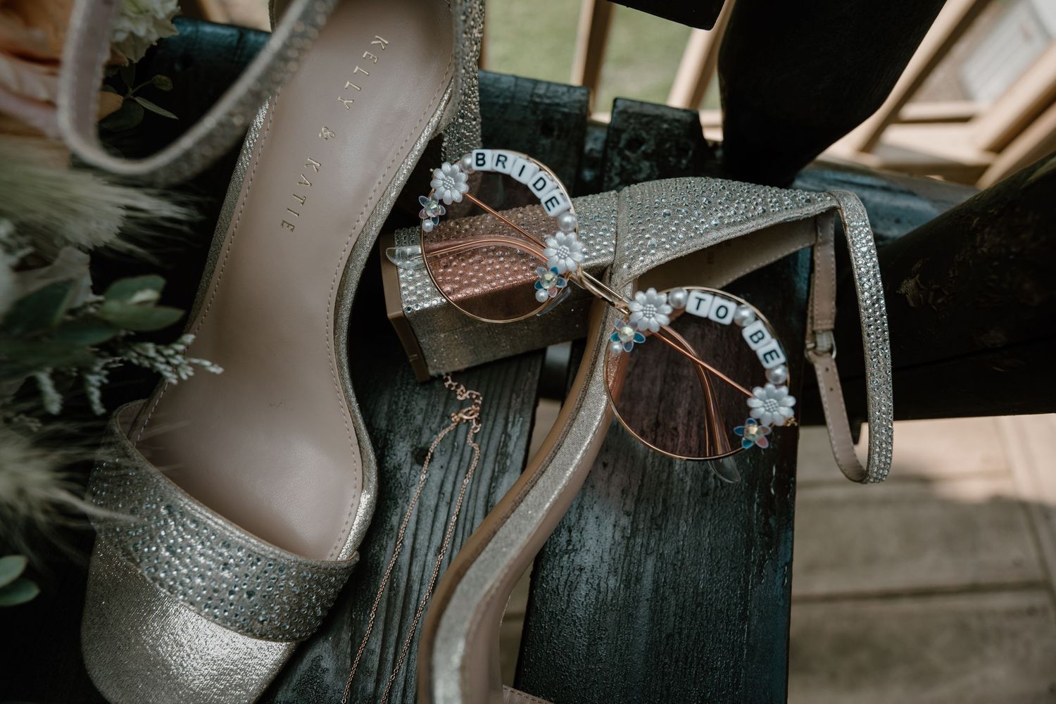 Silver bridal heels and rhinestone sunglasses reading bride displayed on rustic wooden surface.