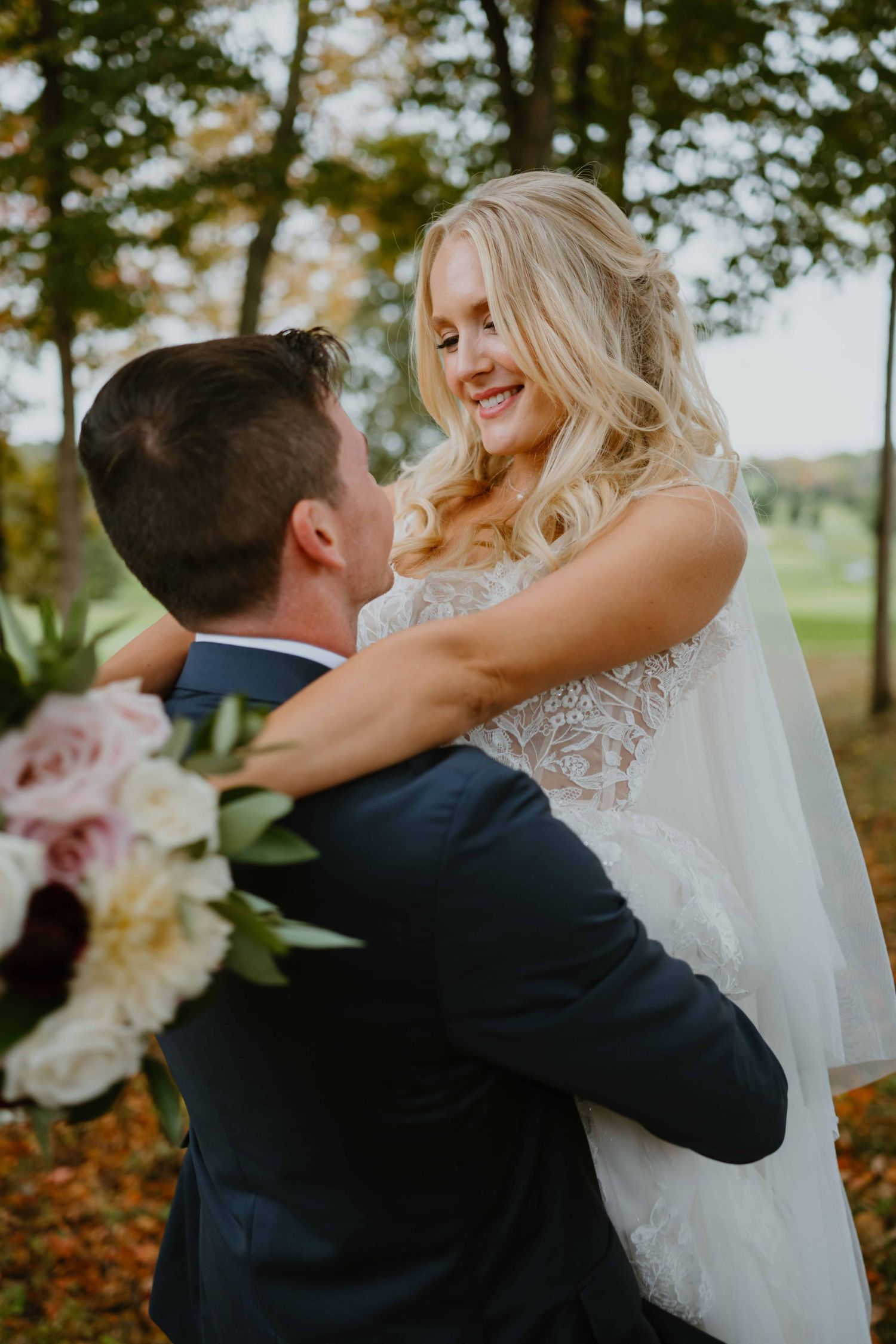 Groom lifting beaming blonde bride holding floral bouquet in autumn setting.