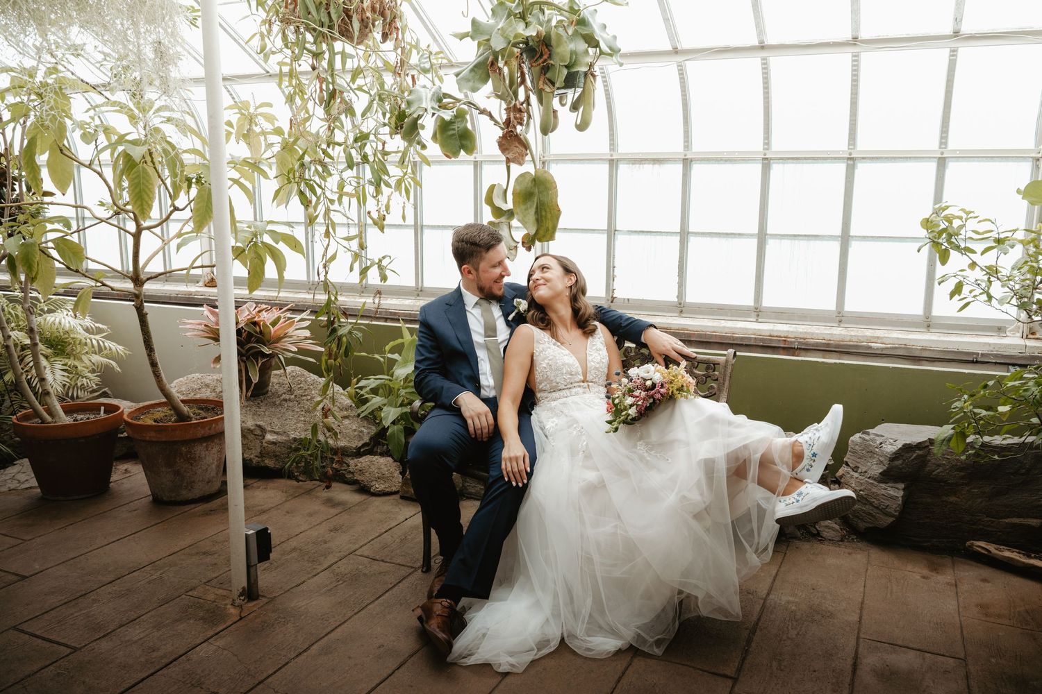 Bride and groom sitting together laughing in a lush greenhouse conservatory on their wedding day.