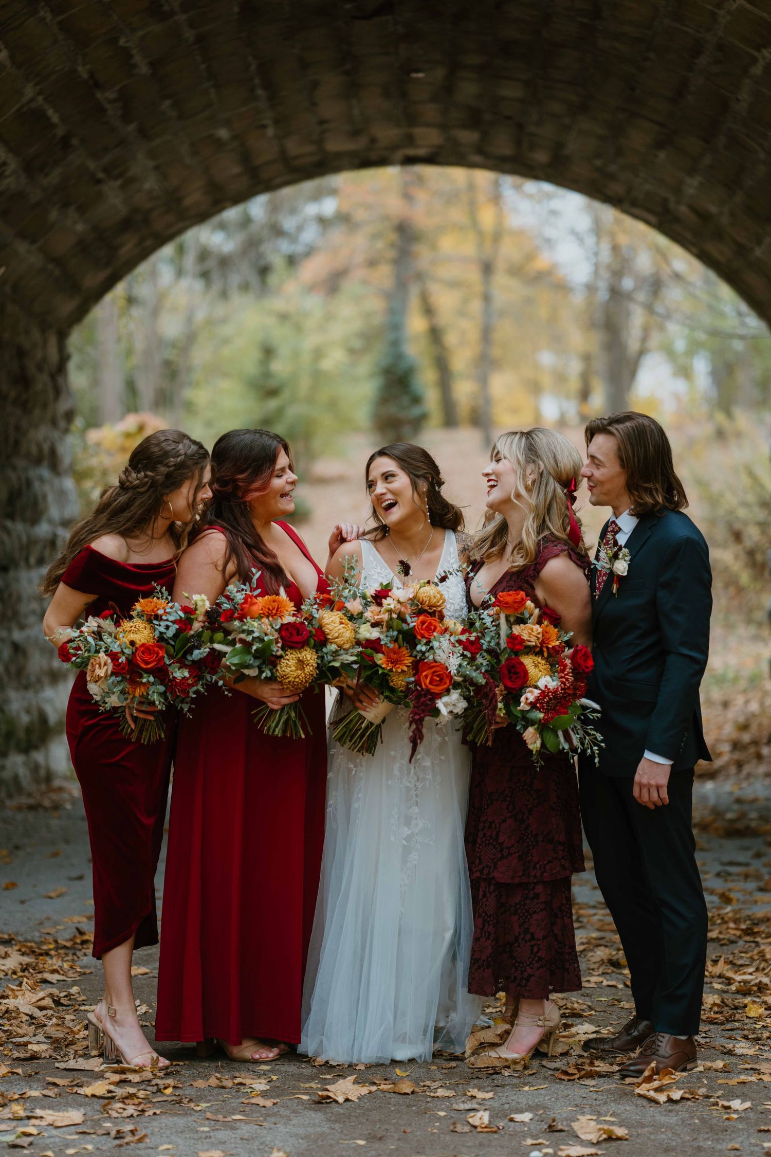 Bridal party laughing together under stone arch bridge surrounded by autumn foliage and red bouquets.