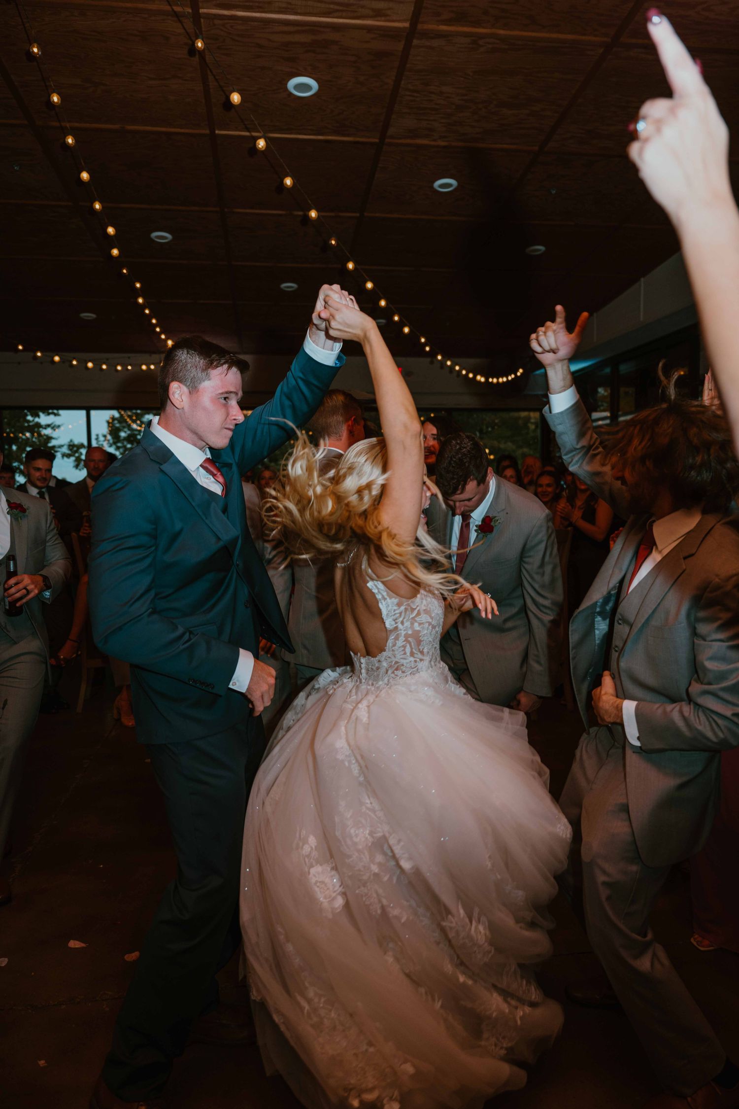 Bride and groom dancing joyfully at wedding reception with string lights and guests surrounding them.