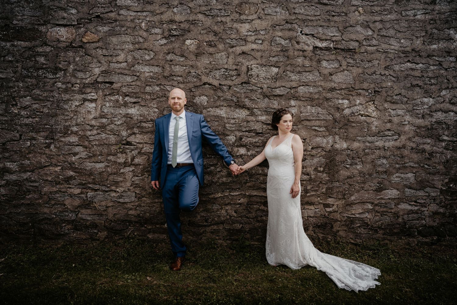 Bride and groom hold hands laughing together against rustic stone wall outdoor wedding portrait.