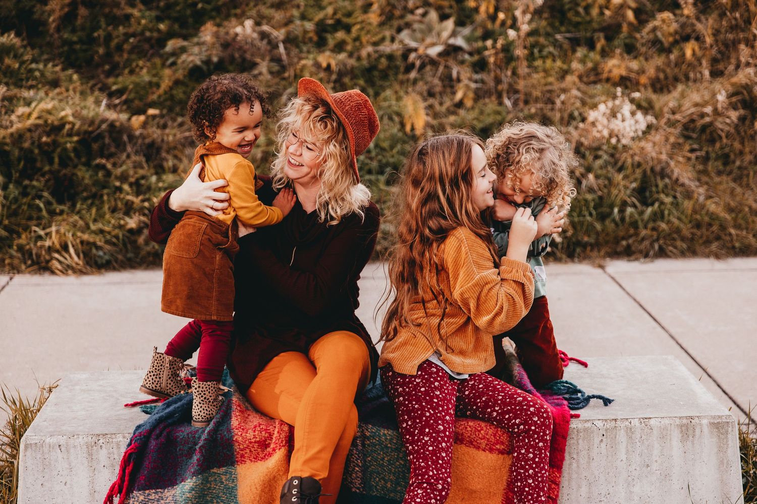 A family laughs together as they sit on a concrete bench with foliage in the background.