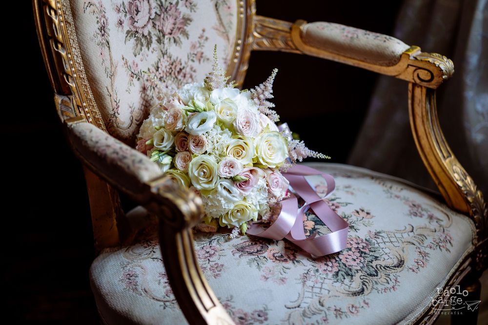 A bridal bouquet of white roses and wedding shoes rest on an antique floral chair.