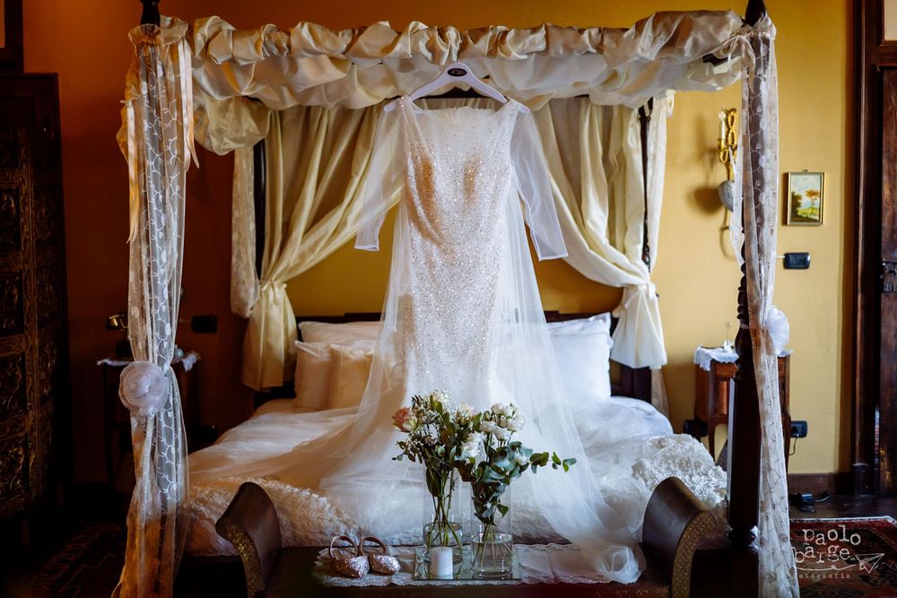 White lace wedding dress displayed on elegant canopy bed frame with sheer curtains and floral arrangement.