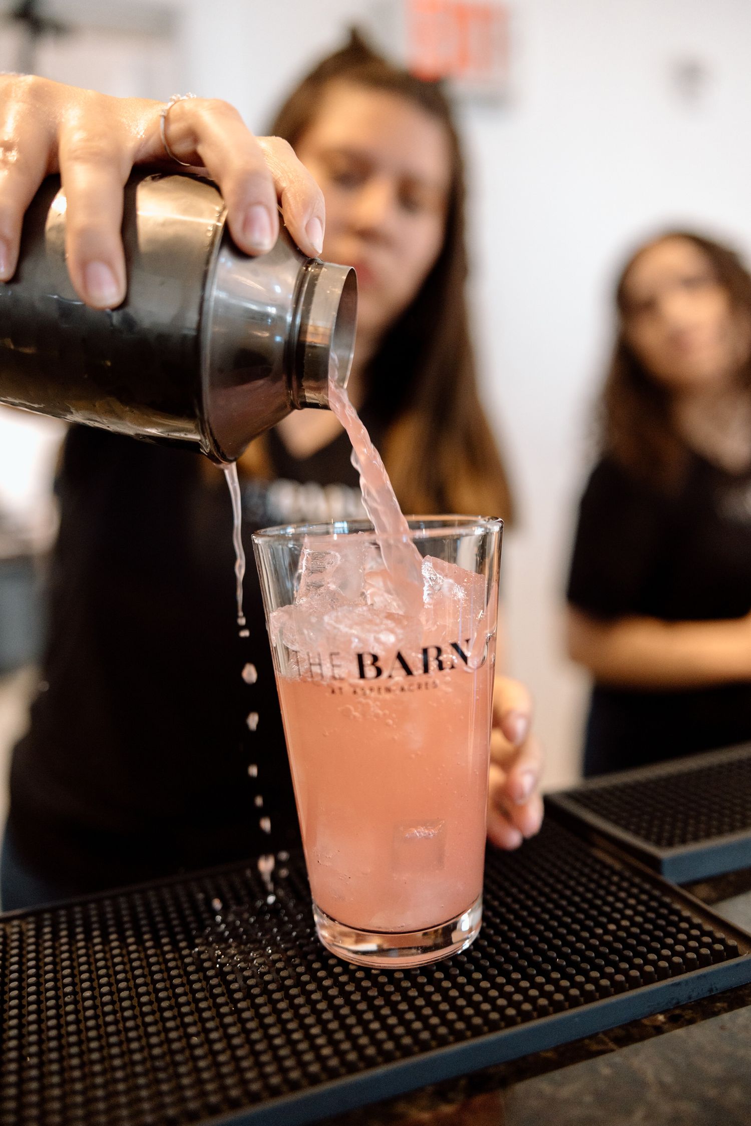 Pink cocktail being poured at a bar with ice and glassware.