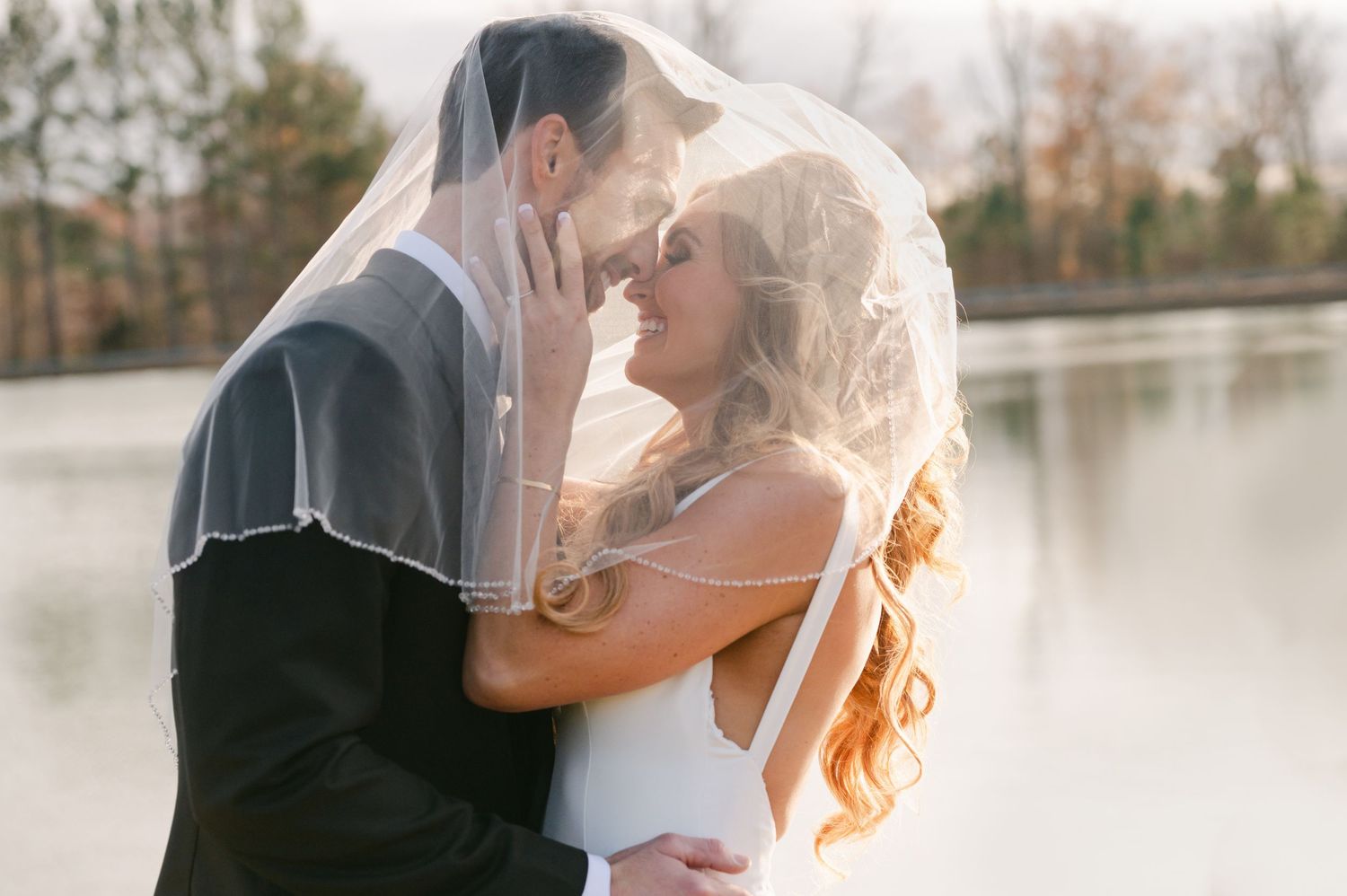 Romantic bride and groom share an intimate moment under a veil by a shimmering lake at sunset.
