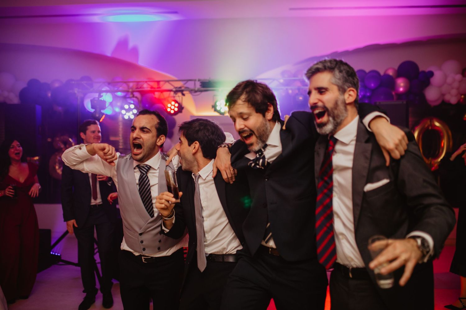 Wedding party dancing and celebrating under vibrant purple and pink lighting at reception.