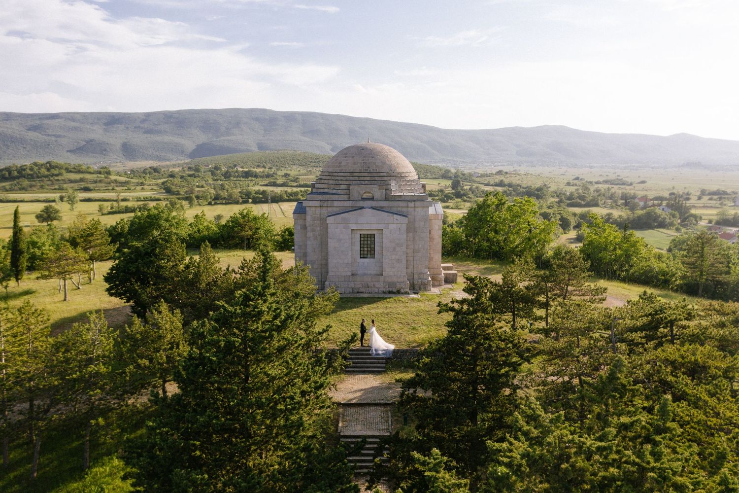 A small domed stone building stands on a hilltop overlooking a green rural landscape with mountains in the distance.