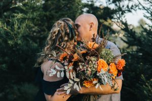 A romantic couple embraces while holding a bouquet of vibrant orange flowers against a natural outdoor backdrop.