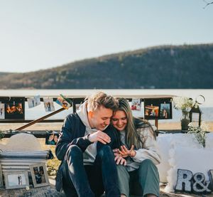 Two people share a tender moment while looking at something together on a bench overlooking mountains.
