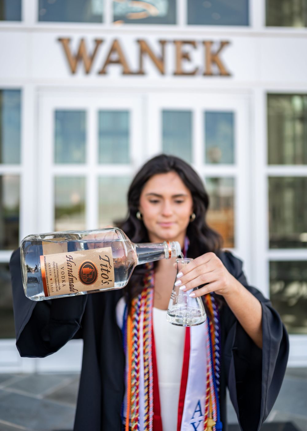 Graduate in academic regalia pouring celebratory drink outside Wanek building.