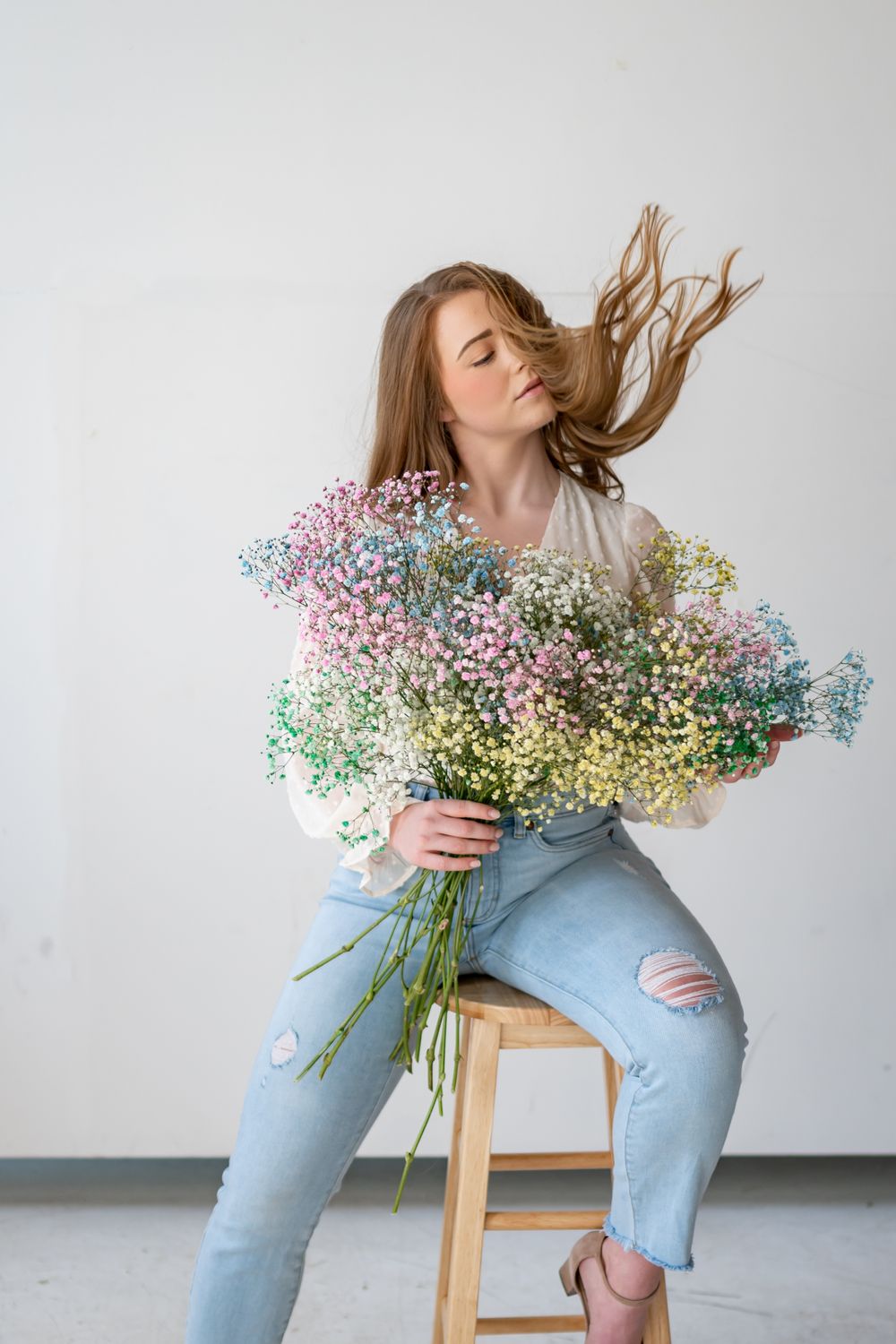 Model holding colorful baby's breath bouquet while sitting on wooden stool.