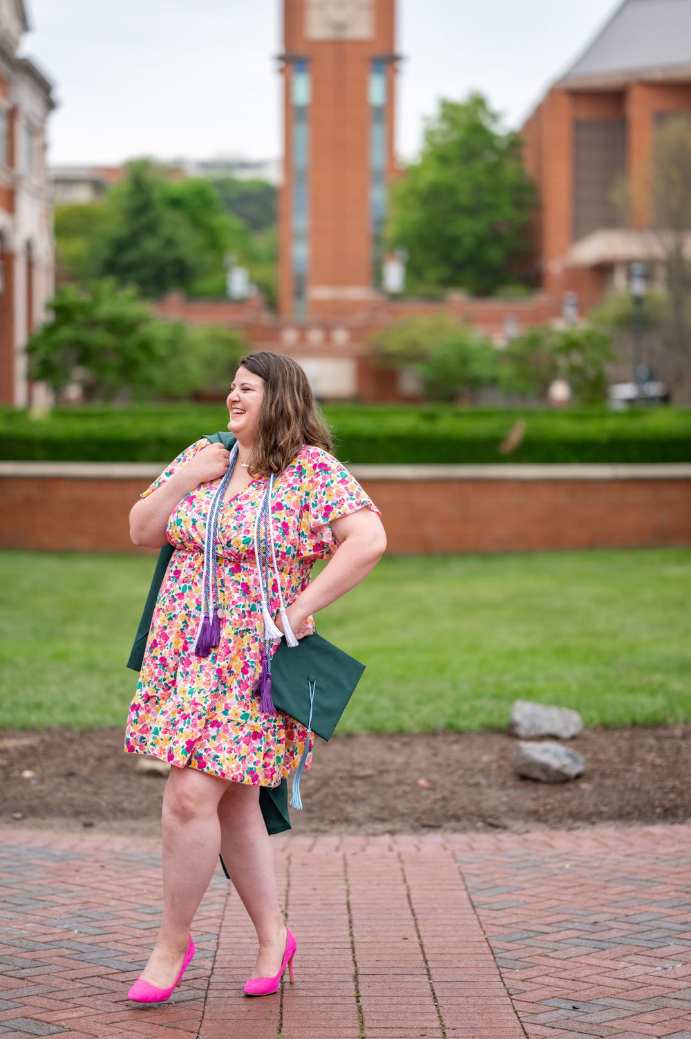Graduate in floral dress and green cap posing on brick pathway near university building.