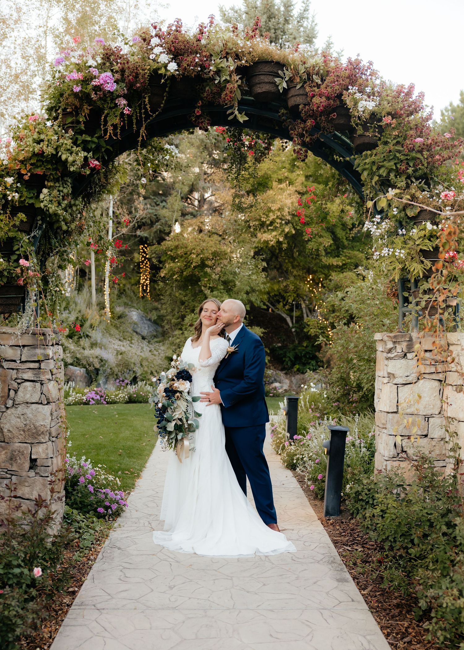 Groom kissing brides cheek under floral arch at Thanksgiving Point Gardens in Lehi, Utah