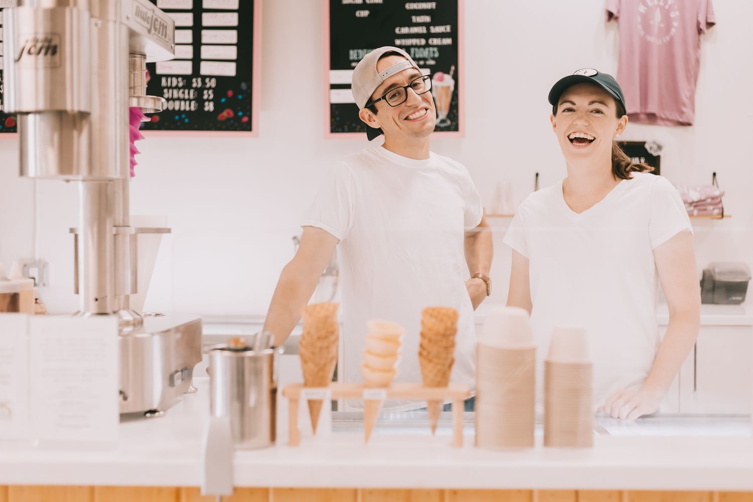A couple of ice cream shop owners stand behind counter smiling. Cones and cups on counter