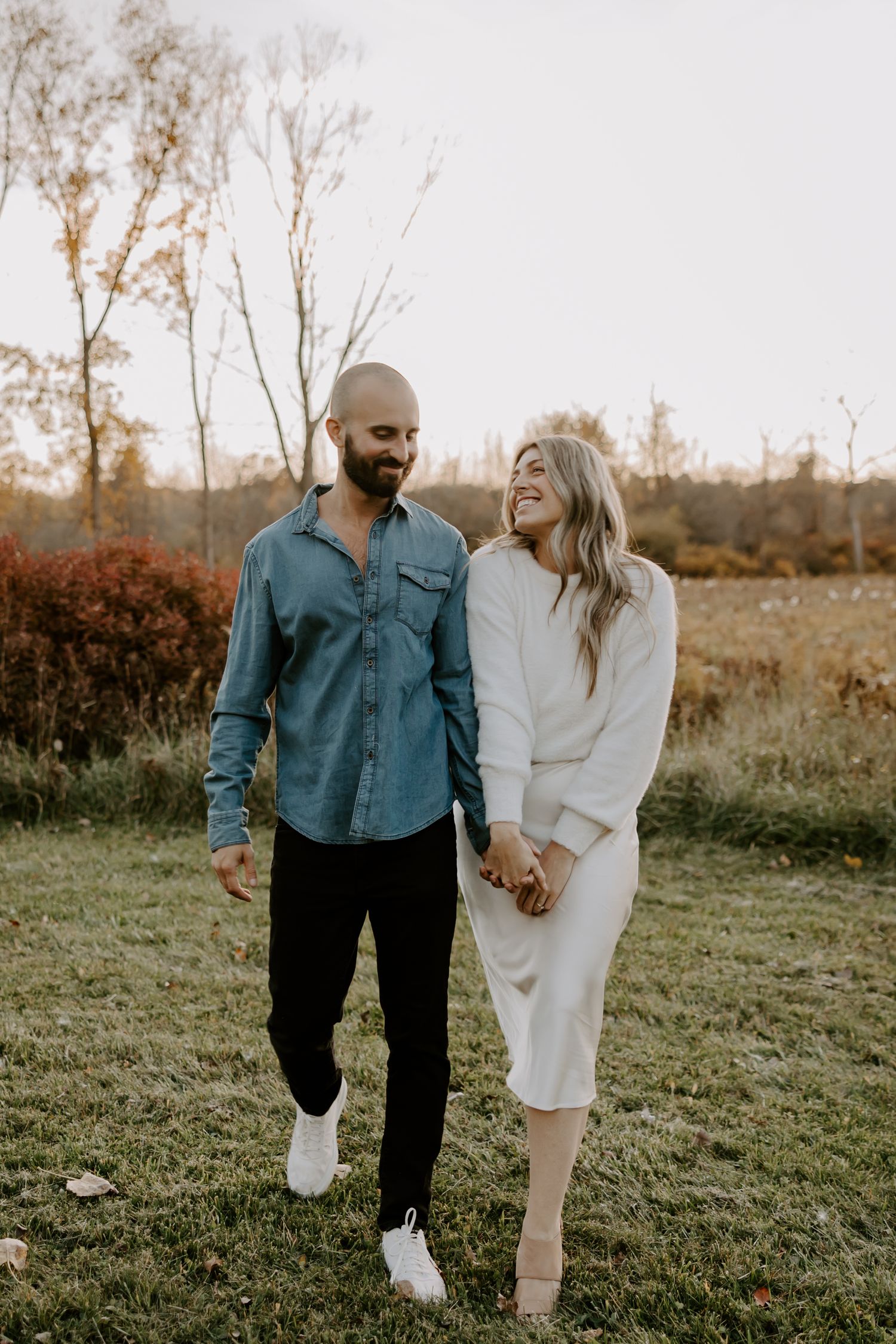 Happy couple holding hands and walking through an autumn field at golden hour, man in blue shirt and woman in white dress.