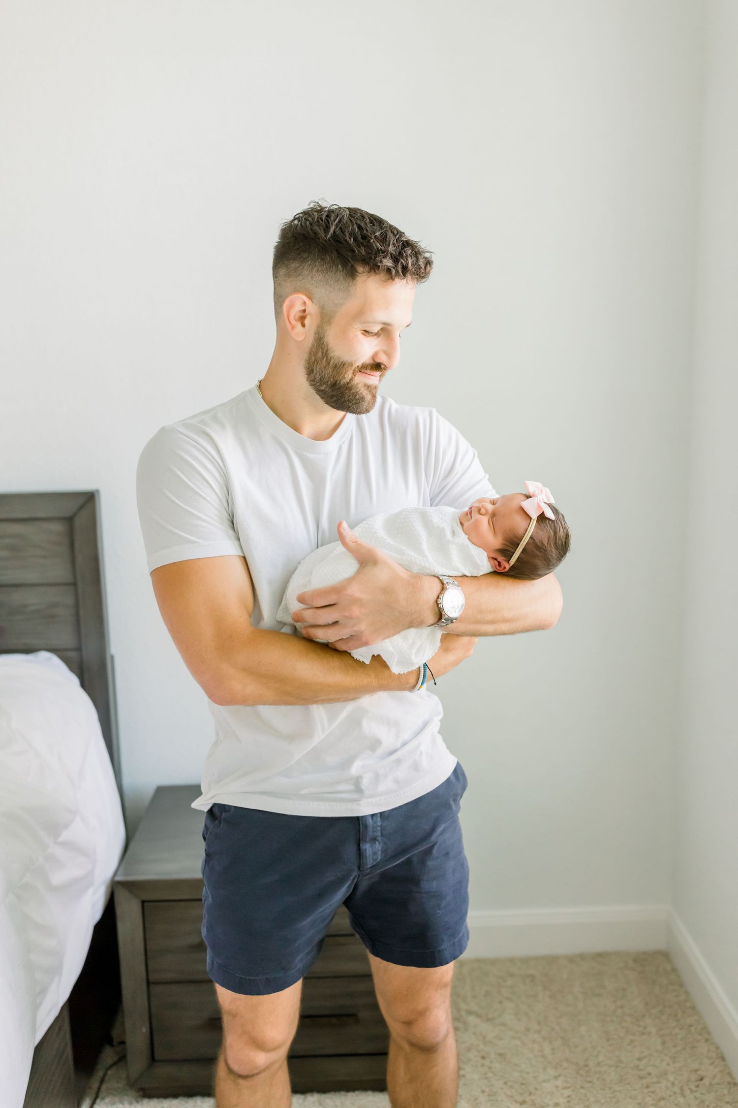An adult holds a baby while standing beside a gray headboard bed.