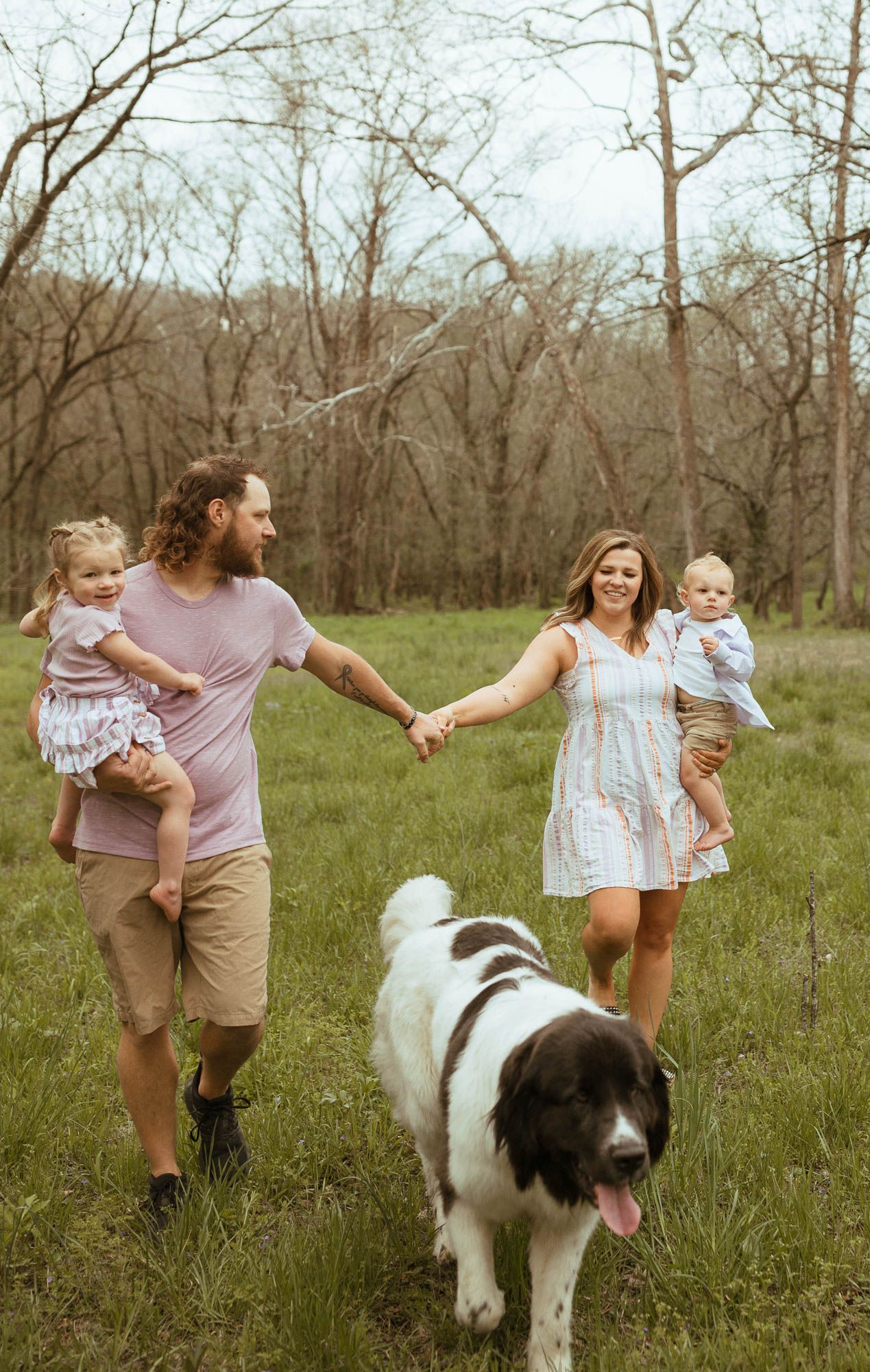 A family walks through a spring meadow holding hands with their large fluffy dog.
