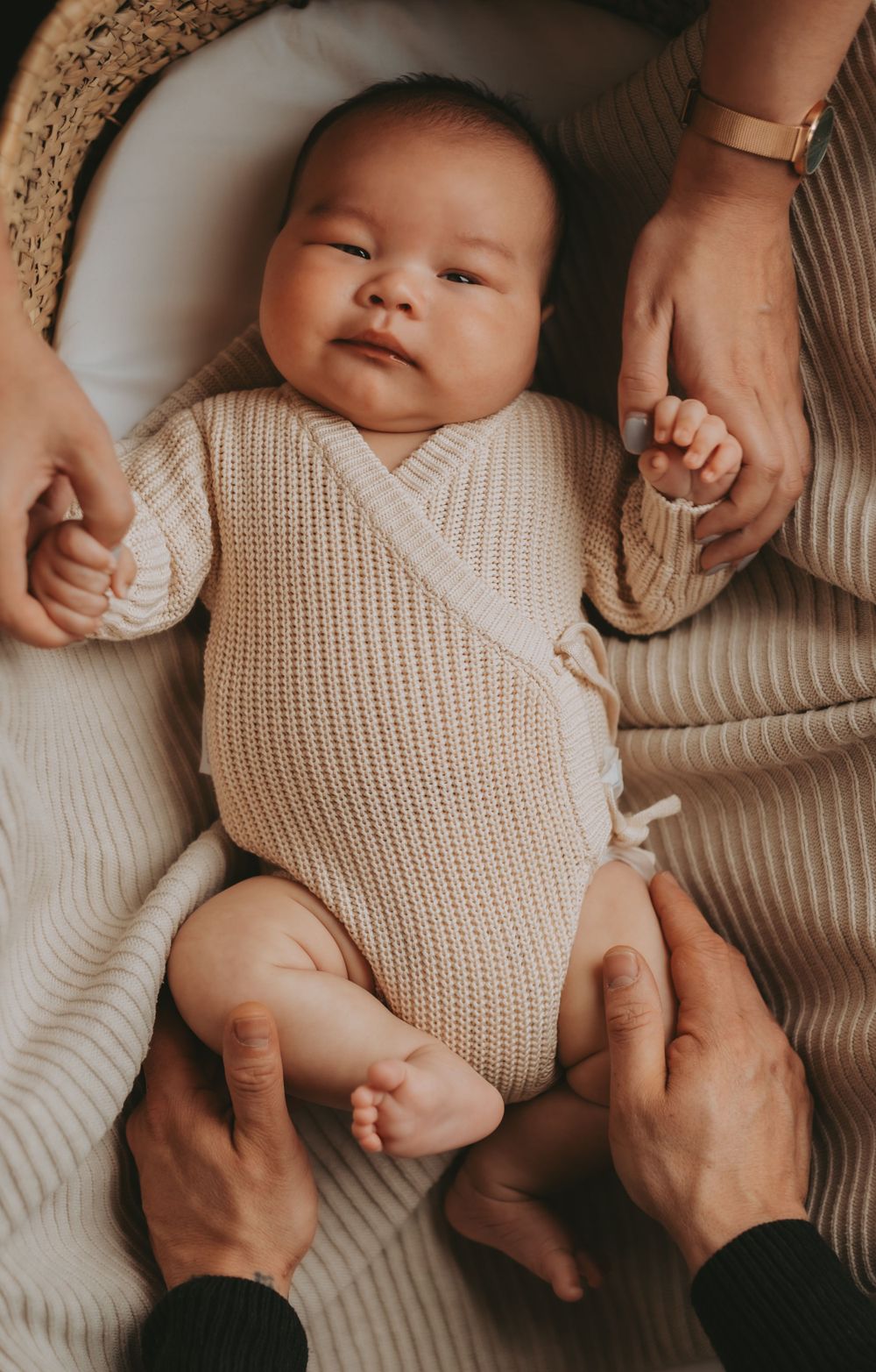 A baby wearing a cream knit sweater lies comfortably while being held by caring hands.