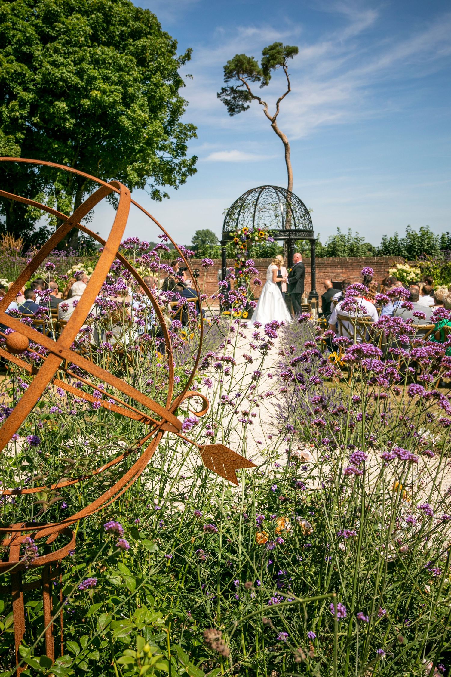 Garden wedding venue with metal gazebo and purple flowers against blue sky.