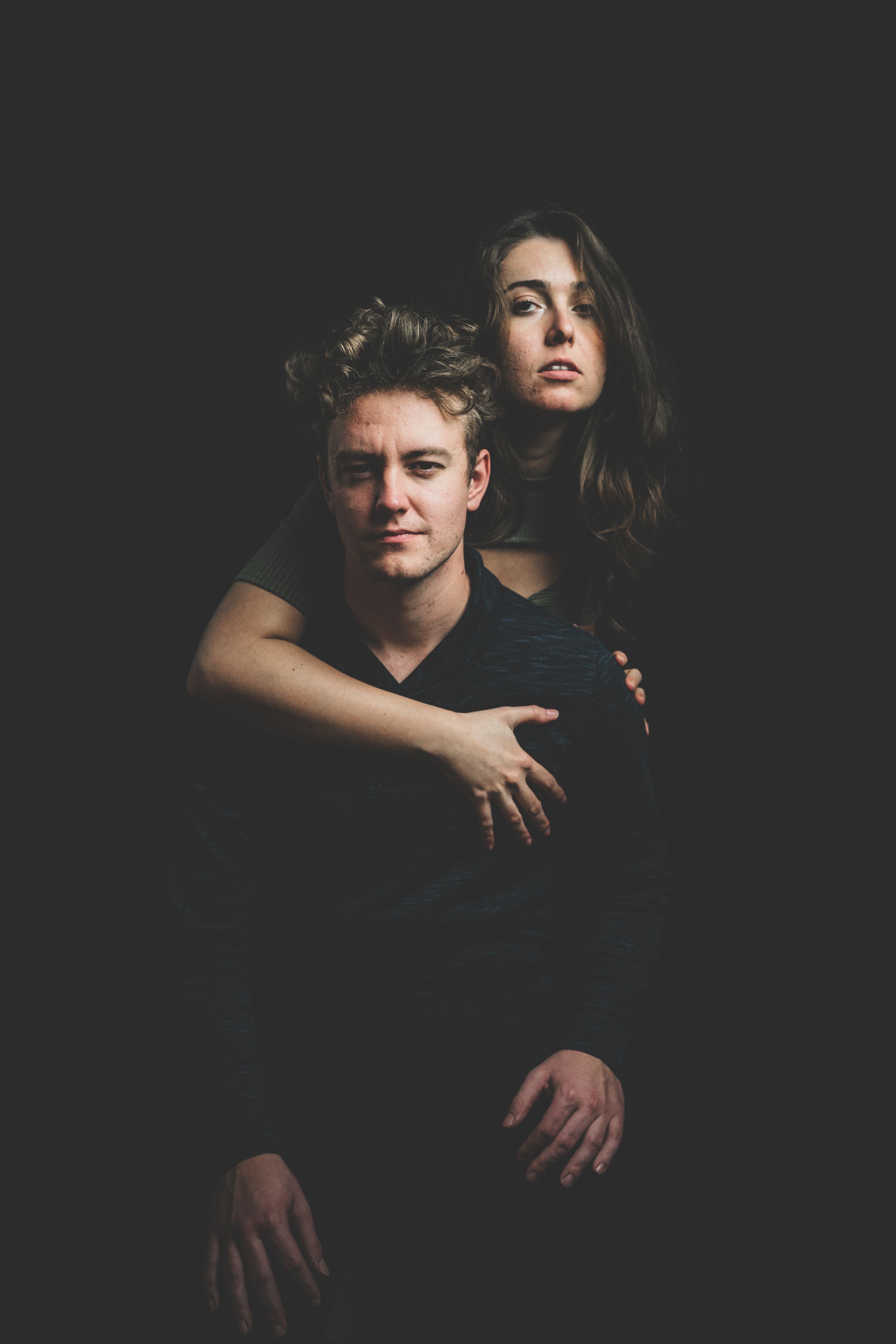 Couple posing dramatically in dark studio, woman embracing man from behind on black background.