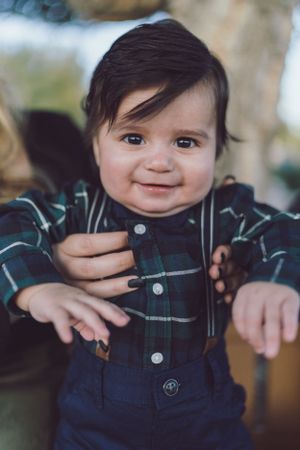 An adorable smiling baby boy in a plaid shirt with suspenders reaches toward the camera outdoors.