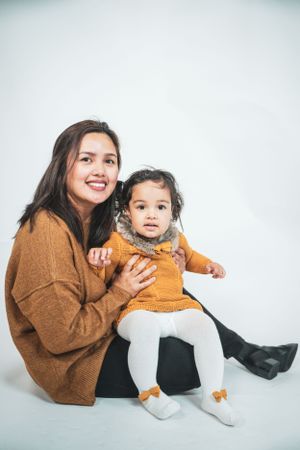 A smiling mother in an orange sweater sits with her baby daughter dressed in matching mustard outfit.
