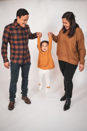 Smiling parents lift their toddler daughter by the hands against a white studio background.