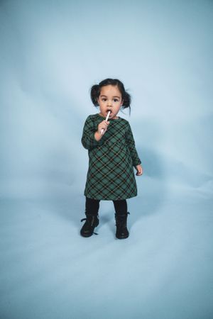 An adorable toddler girl in a green plaid dress and black boots holds a lollipop on blue background.