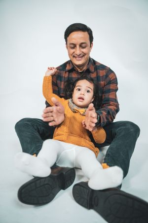 A happy father sits on the floor playing with his toddler daughter in a white studio setting.