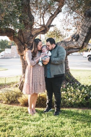 Young family with newborn baby kissing under a large tree during outdoor photo session.