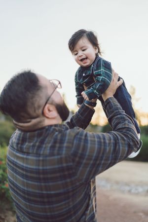 Joyful father lifting laughing baby in the air during outdoor family photo session.