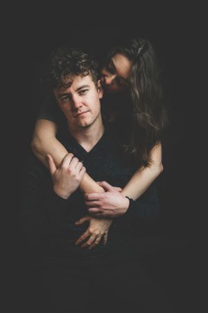 A woman wraps her arms around a man from behind in a dark, intimate studio portrait.