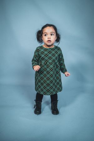 A curious toddler girl in a green plaid dress and black boots stands on a blue background.