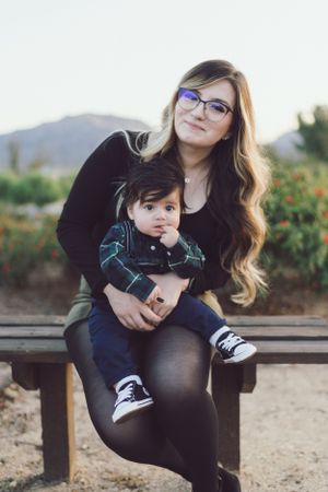 Young mother with glasses sitting on bench holding baby during outdoor portrait session.